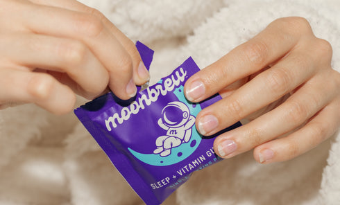 Close-up of hands opening a MoonBrew Sleep + Vitamin Gummies single-serve purple packet on a beige blanket