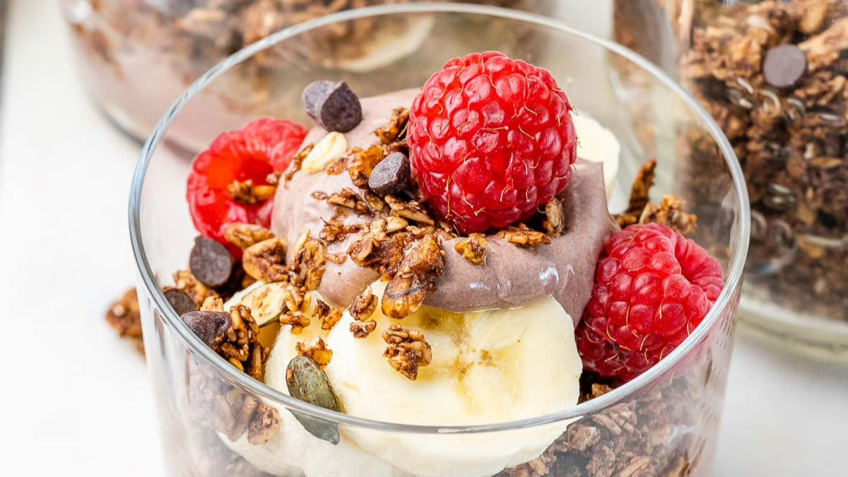 Chocolate yogurt parfait layered with banana slices, granola, chocolate chips, and raspberries in a clear glass