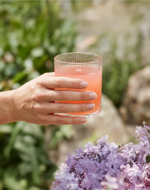 Hand holding a ribbed glass of pink sleep support drink above lilac flowers in a garden setting