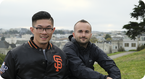 Two men sitting on a grassy hill in a park with a city skyline behind them, one wearing a black jacket with SF logo