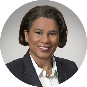 Professional headshot of a woman wearing a dark blazer and white blouse against a neutral gray vignette background