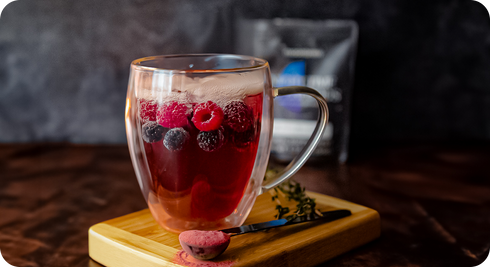 Glass mug of hot berry infusion with raspberries and blueberries on wood board, spoon with pink powder and blurred pouch