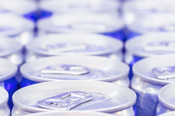 Close-up of energy drink can tops showing pull tabs, cool blue lighting, and blurred rows of cans in the background