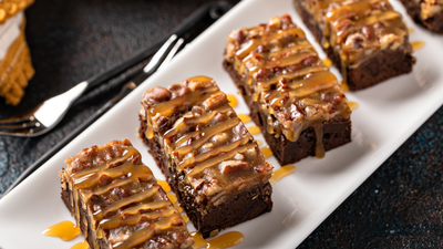 Close-up of caramel-drizzled chocolate bars with pecan topping and fudgy brownie base on a white rectangular plate