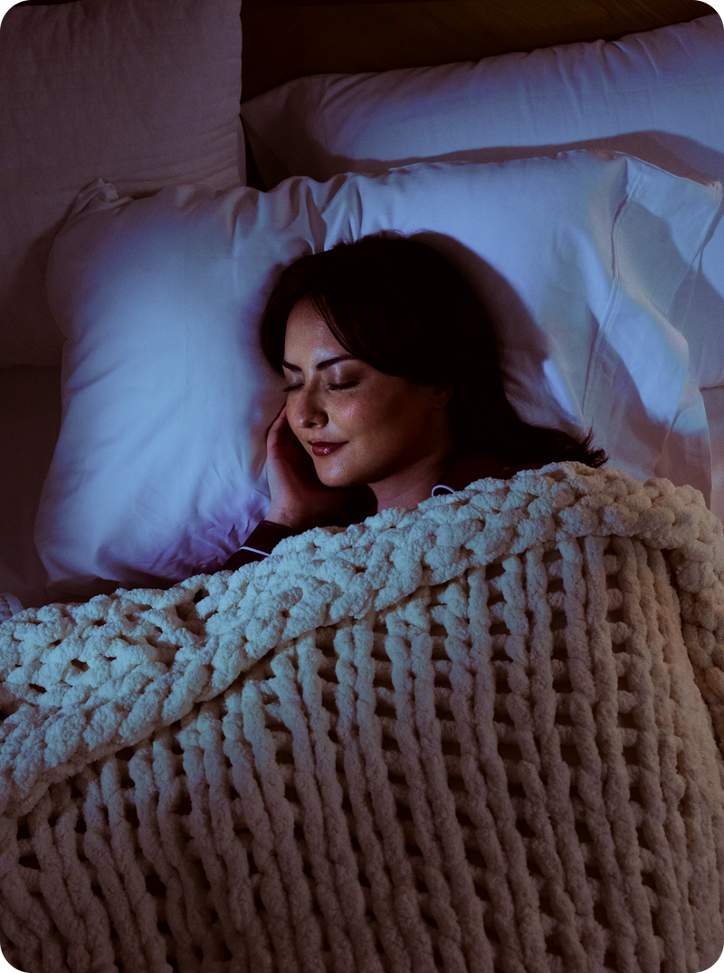 Woman sleeping on her side with earbuds, covered by a chunky cream knit blanket on a pillow in dim bedroom lighting
