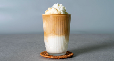 Front view of a vanilla frappe in a tall glass showcasing layered milk and coffee and whipped cream on top against a soft gray background
