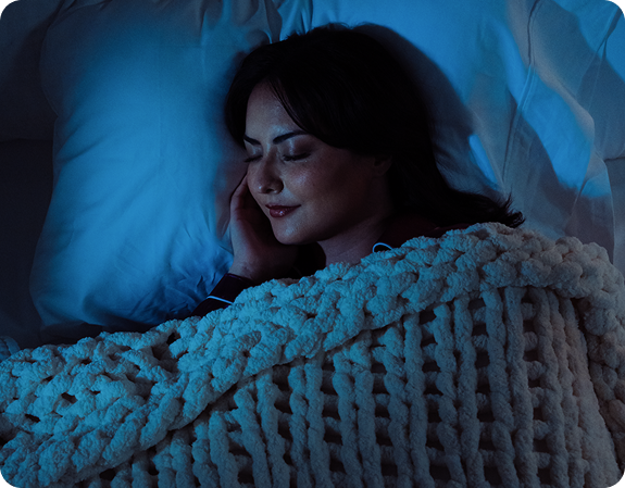 Woman sleeping peacefully on her side under a chunky knit blanket in dim blue bedroom lighting