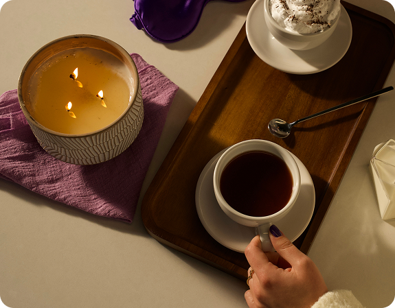 Hand reaching for a white teacup on a wooden tray beside a lit three-wick candle on a purple cloth, cozy nighttime setting