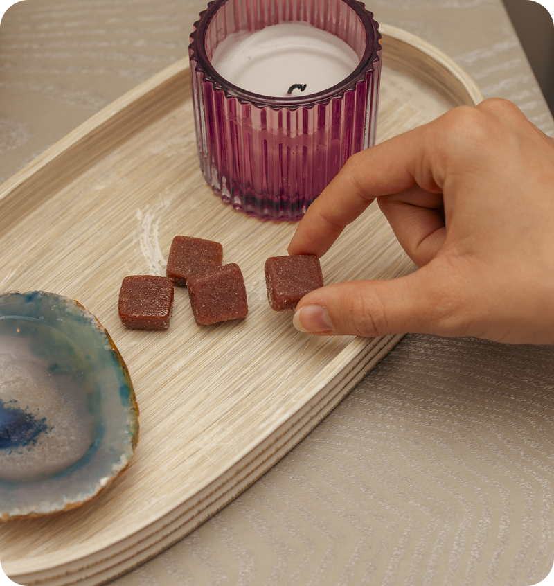 Hand picking a square reddish brown gummy from a wooden tray beside a purple glass candle and coaster