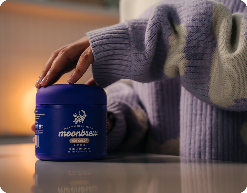Close-up front view of MoonBrew Hot Cocoa jar showcasing product packaging and hand resting on the lid with a blurred warm indoor background