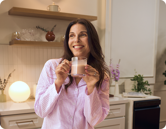 Woman in pink pajamas holding a glass mug of warm MoonBrew drink in a lit kitchen, smiling