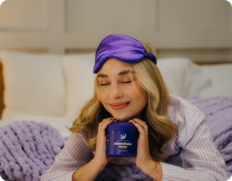 Woman smiling with eyes closed holding a purple MoonBrew jar on a bed, wearing a purple silk sleep mask and striped pajamas