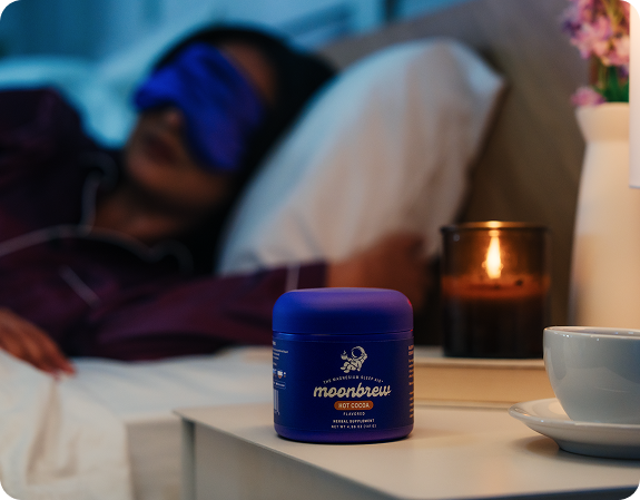 MoonBrew jar on bedside table with a cup and lit candle, blurred person sleeping in bed in the background