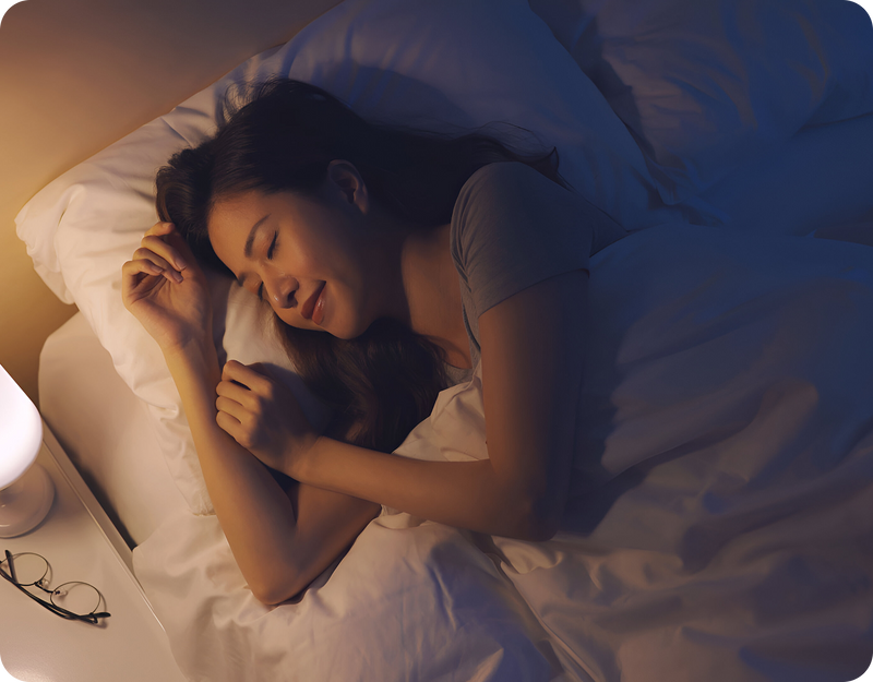 Woman sleeping peacefully in bed at night with a warm bedside lamp and glasses on the nightstand