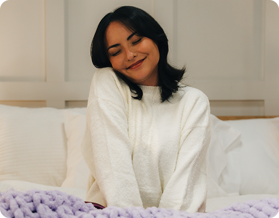 Woman sitting on a bed, eyes closed and smiling, wearing a white fuzzy sweater with a purple knit blanket, relaxed bedroom