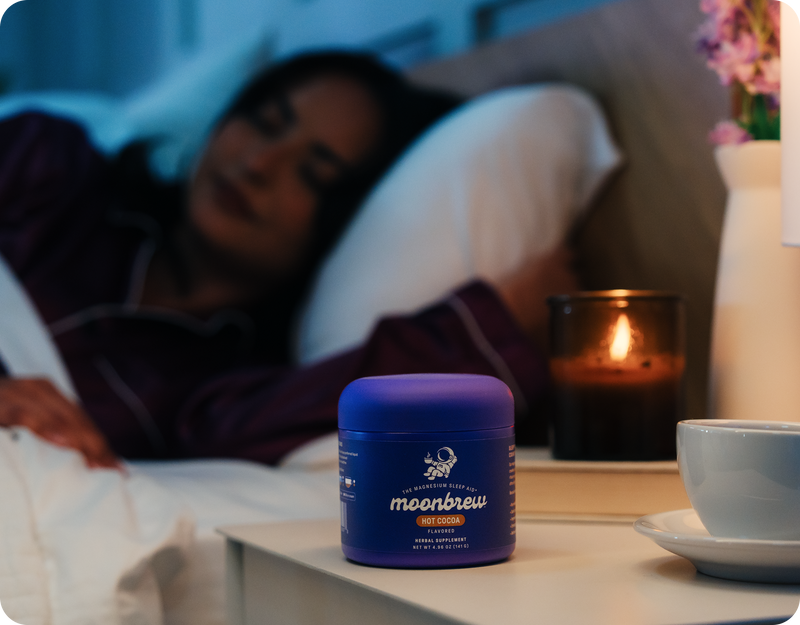 MoonBrew jar on a bedside table beside a lit candle and teacup with a person sleeping in the background