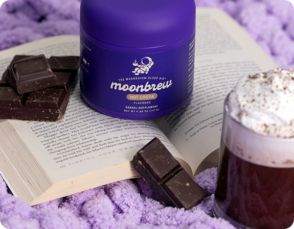 Angled front view of MoonBrew Hot Cocoa tub with chocolate squares and a whipped-cream mug on a book and purple blanket