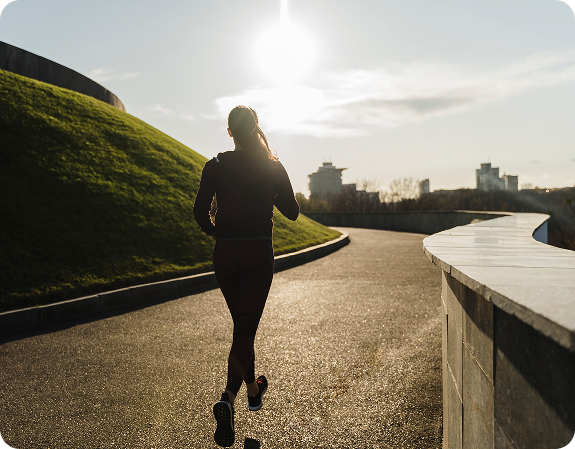 Behind angle of a person jogging on a sunlit park pathway at sunrise with city skyline in the distance