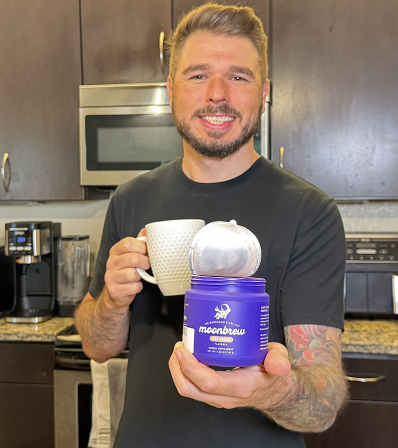 Smiling man holding an open purple Moonbrew jar labeled moonbrew and a white mug in a kitchen