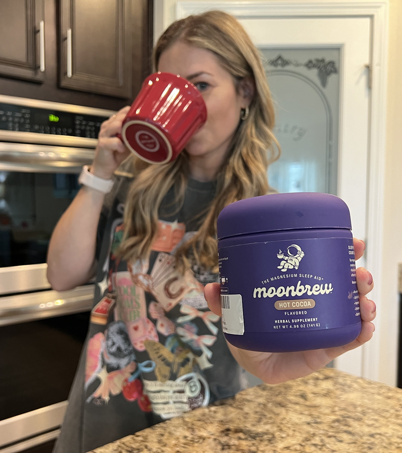 Front view of MoonBrew Hot Cocoa jar showing purple packaging and visible label held by a person in a kitchen with a blurred background