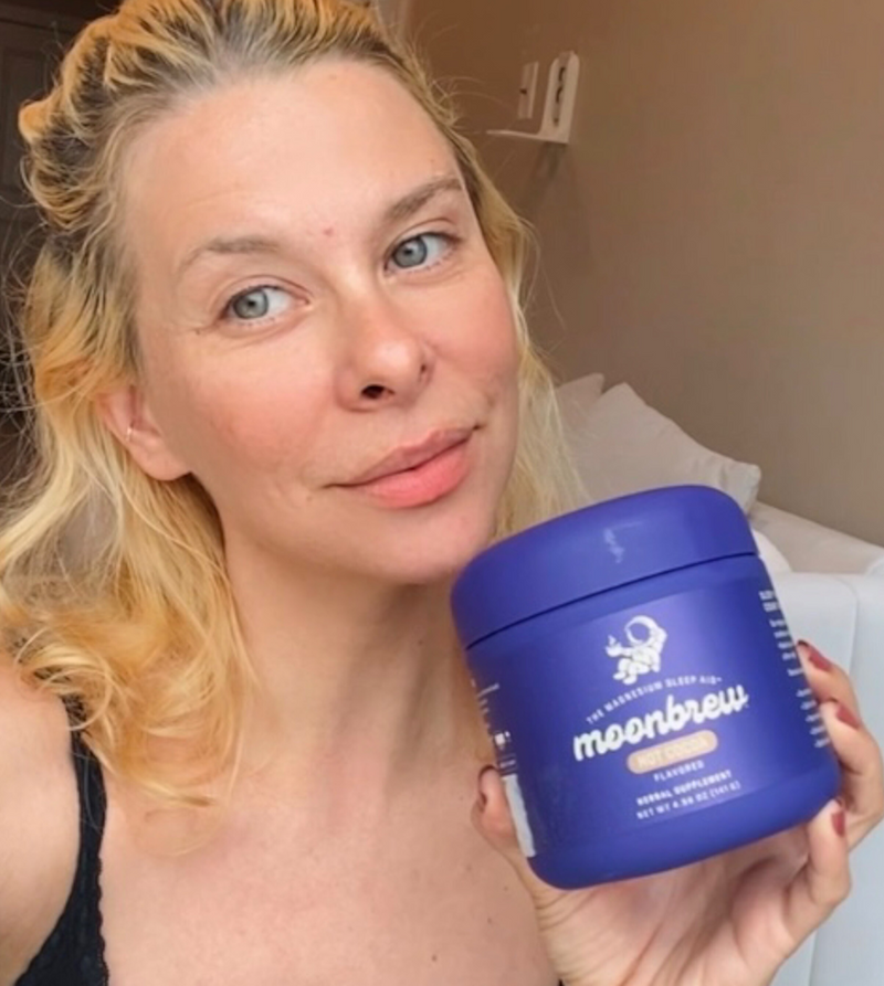 Person holding a purple MoonBrew supplement jar near their face in a bedroom setting with the product label visible