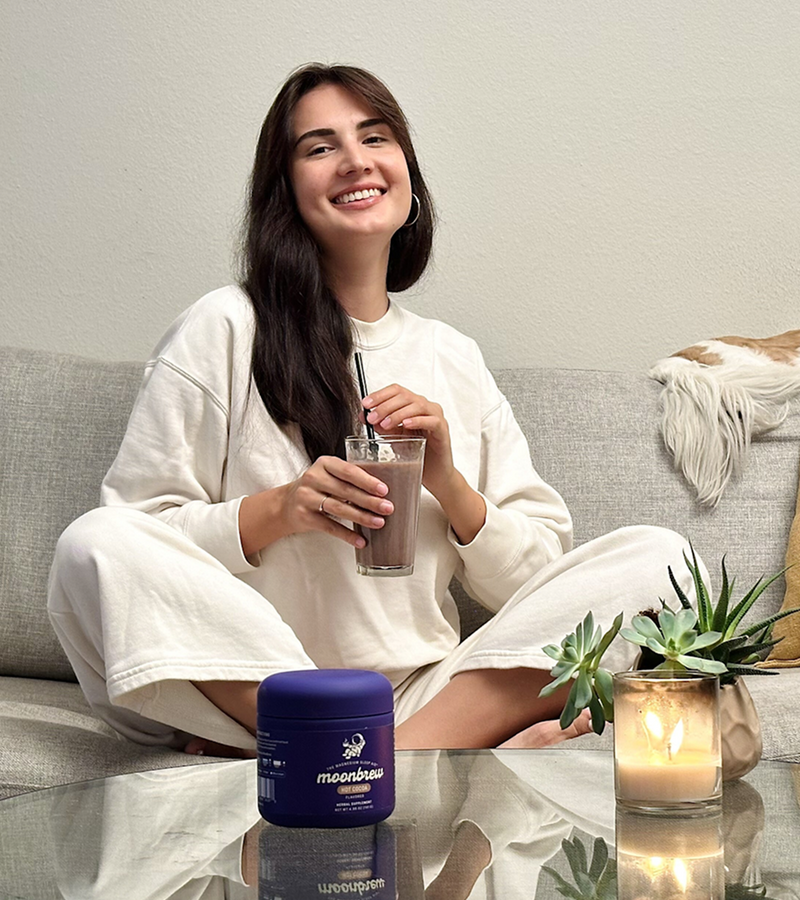 Woman on a couch sipping a chocolate drink with a MoonBrew tub on the coffee table and a lit candle