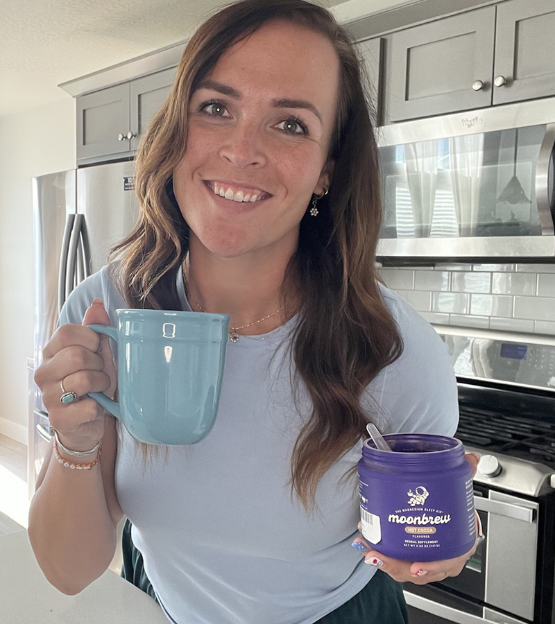 Woman smiling holding a blue mug and a purple MoonBrew jar with a spoon in a bright kitchen