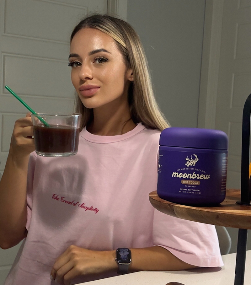 Woman holding a glass mug of hot cocoa beside a purple MoonBrew Hot Cocoa container on a kitchen counter