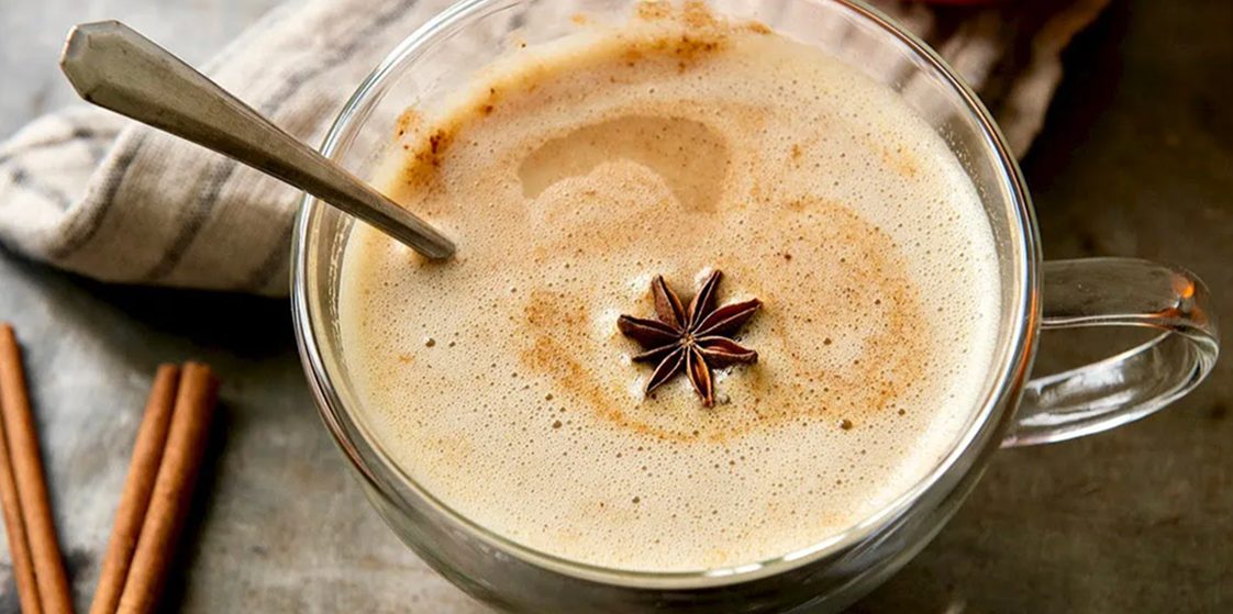 Frothy spiced latte in clear glass mug topped with star anise and cinnamon dust, metal spoon at rim