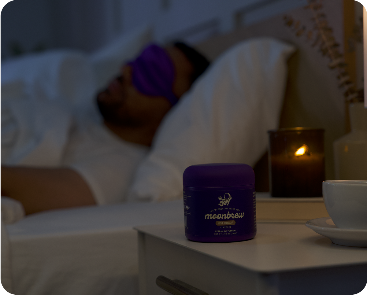 Person sleeping with purple sleep mask, MoonBrew Hot Cocoa supplement jar on bedside table with candle and cup in dim bedroom
