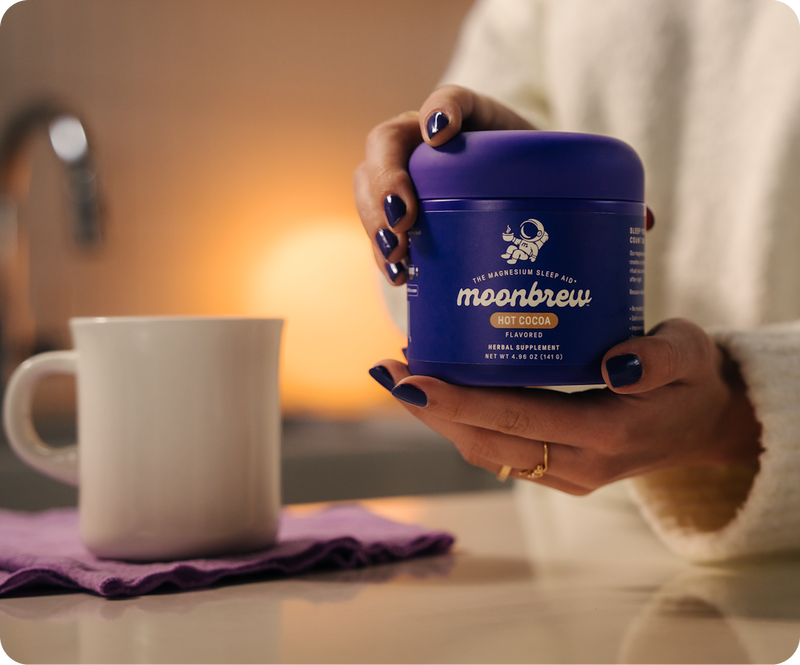 Hands holding a purple MoonBrew Hot Cocoa jar over a kitchen counter next to a white mug with warm background lighting