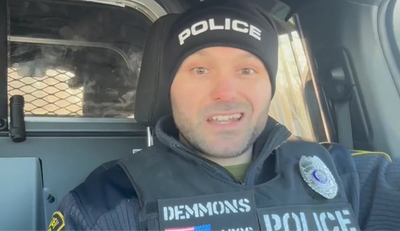 Man wearing a black beanie labeled POLICE and a tactical vest speaking inside a patrol vehicle