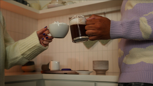 Two people clinking a small foam topped cup and a glass mug of hot cocoa in a cozy kitchen