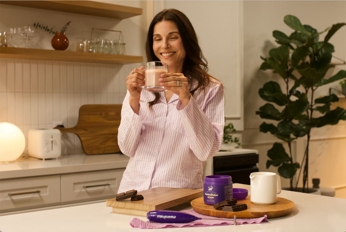 Woman in pink striped pajamas smiling and holding a glass of MoonBrew drink in a kitchen with a MoonBrew canister and chocolate on the counter