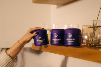 Hand picking a purple MoonBrew jar from a wooden shelf with two other MoonBrew jars and glassware in warm lighting