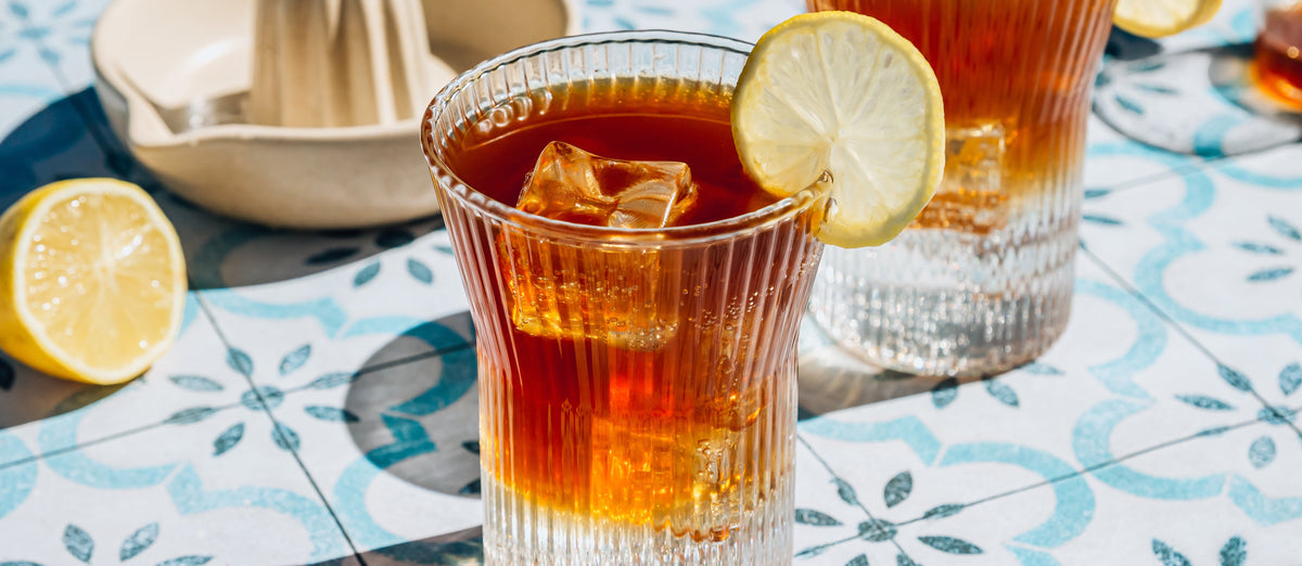 Iced amber recovery drink with lemon slice in a ribbed glass on patterned tile, citrus juicer and halved lemon in background