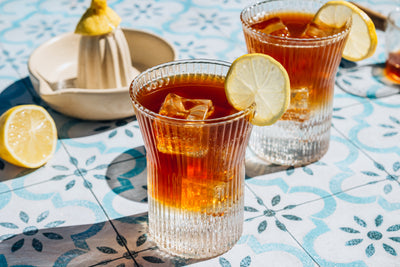 Two iced espresso-tonic drinks garnished with lemon slices on a patterned blue tile table with a citrus juicer nearby