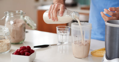Person pouring milk into a shaker to make a chocolate protein shake on a kitchen counter with raspberries and a banana