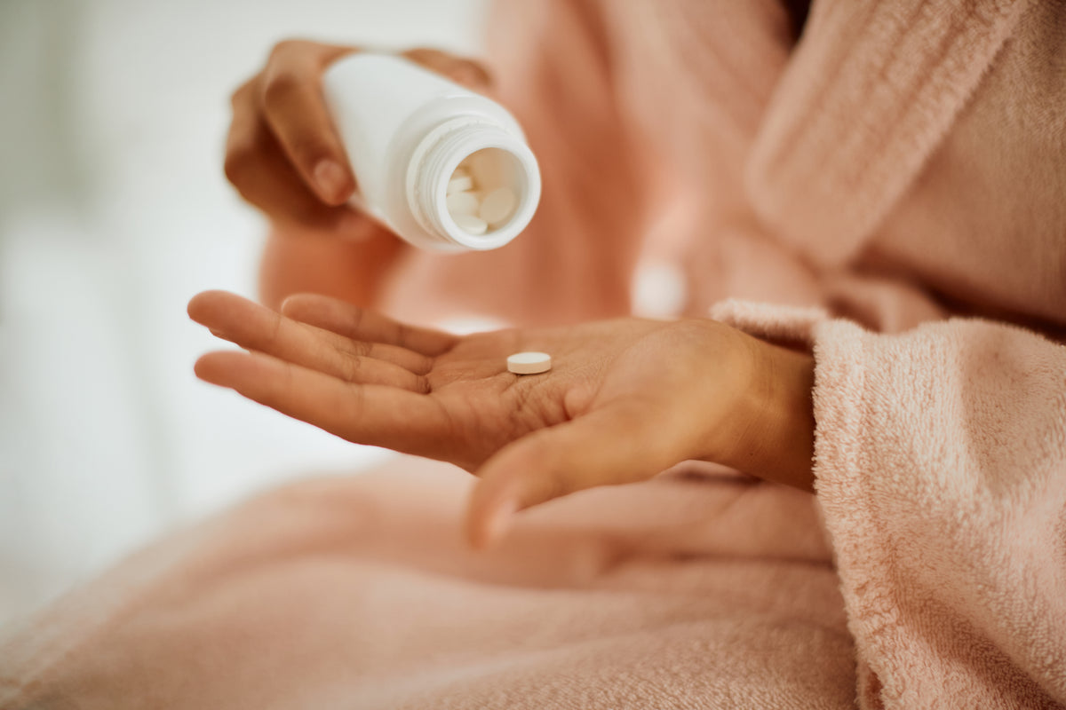 Close-up of a person pouring a white tablet from a pill bottle into their open palm while wearing a pink robe