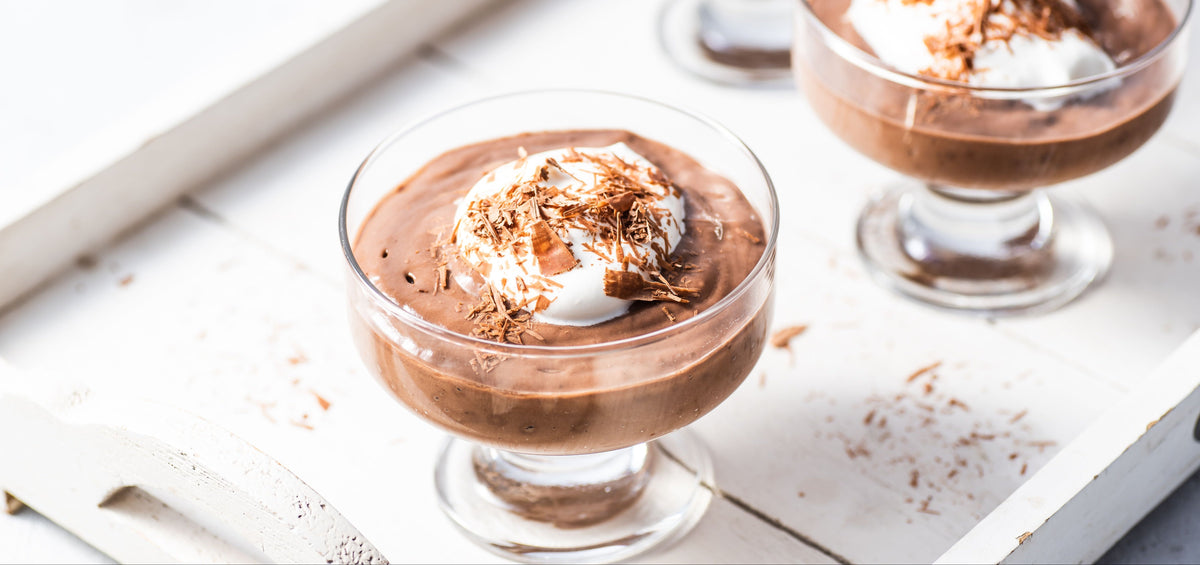 Close-up of chocolate mousse in glass coupe showcasing whipped cream and shaved chocolate on a white wooden tray