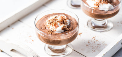Close-up of chocolate mousse in glass coupe showcasing whipped cream and shaved chocolate on a white wooden tray