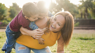Mother laughing as she gives piggyback rides to two young boys in a sunlit park