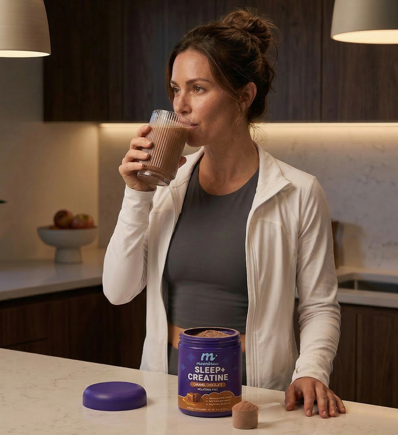 Woman sipping a caramel chocolate MoonBrew Sleep and Creatine shake in a kitchen with open tub and scoop on the counter