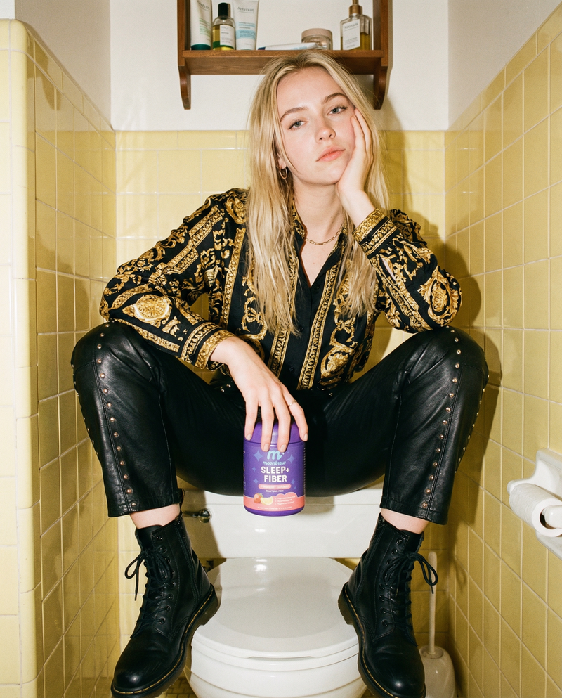 Young woman sitting on a toilet in a yellow-tiled bathroom holding a purple MoonBrew Sleep and Fiber canister