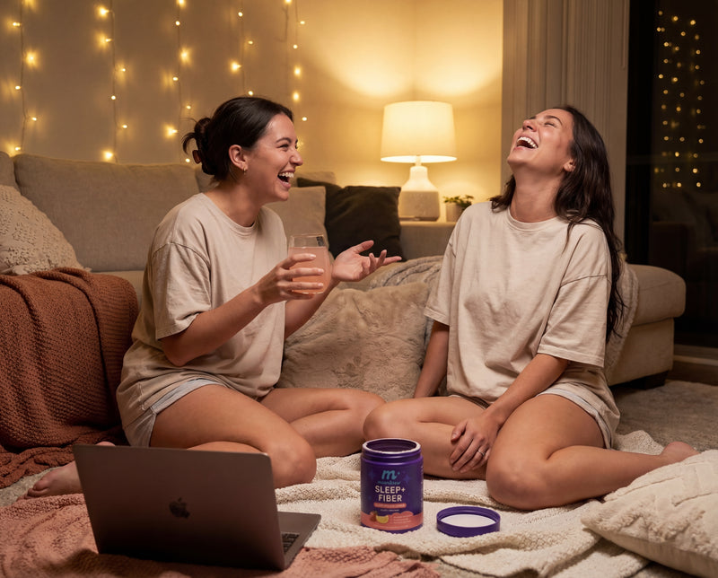 Two women laughing on a living room floor at night with a MoonBrew Sleep+ Fiber jar and glass in foreground, cozy string lights