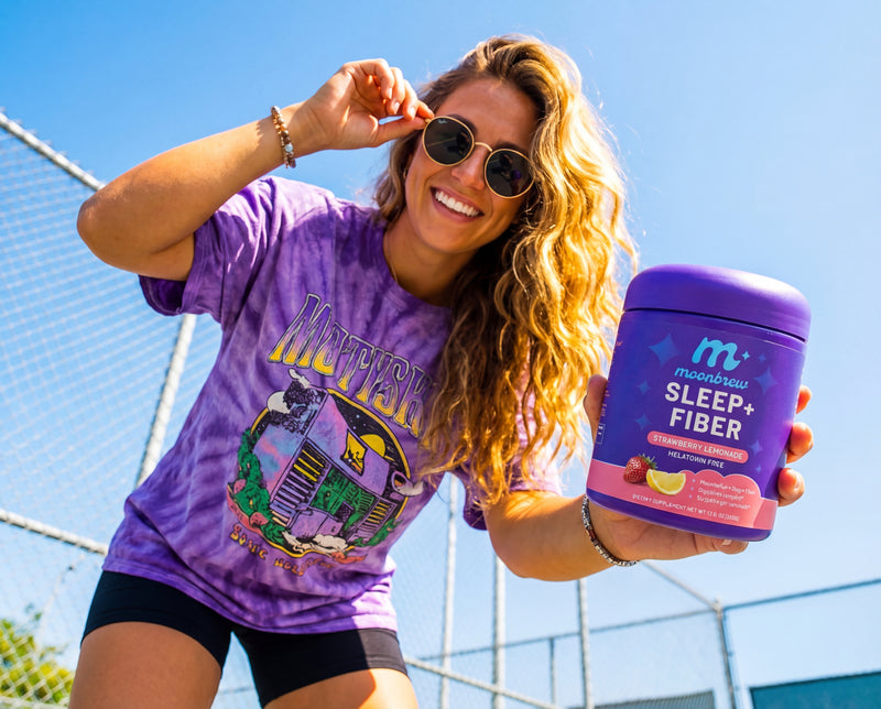 Woman smiling and holding a purple MoonBrew Sleep+Fiber tub (Strawberry Lemonade) toward the camera on a sunny tennis court