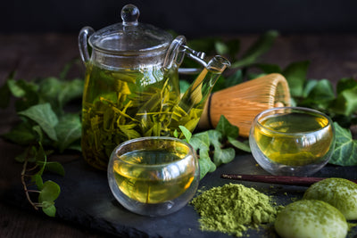 Glass teapot steeping green tea with two glass cups, matcha powder and bamboo whisk on a slate board with green leaves
