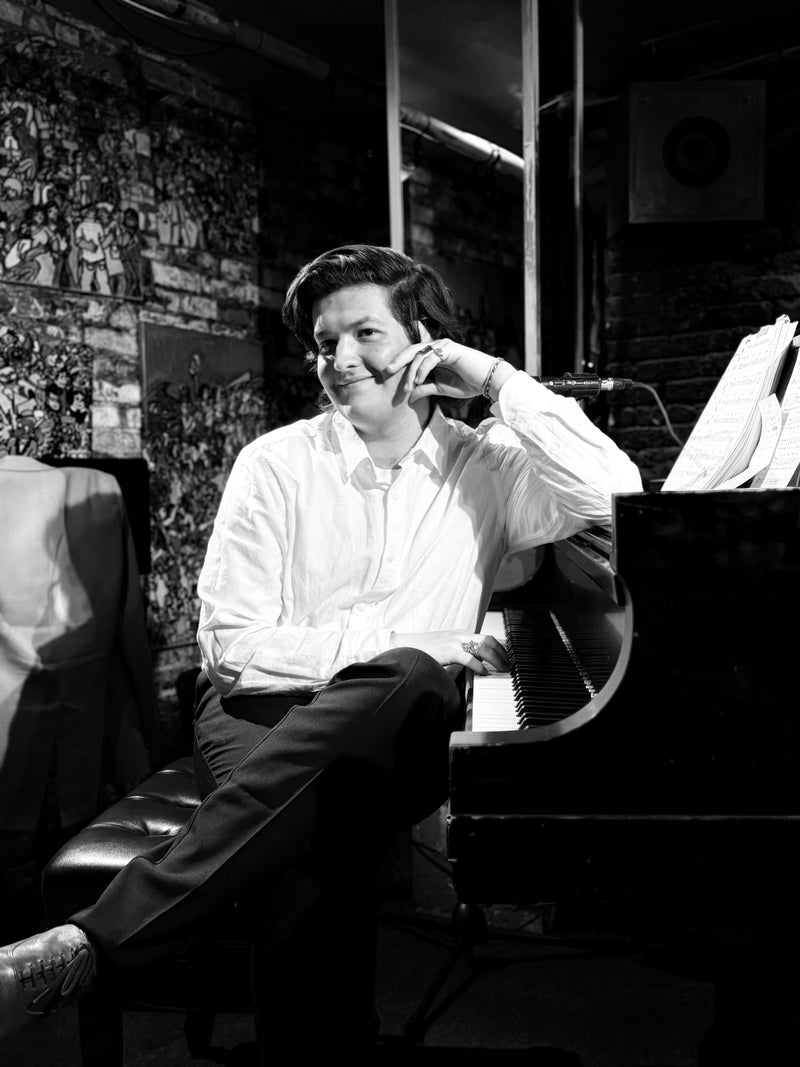 Musician seated at a grand piano smiling mid-set at Smalls Jazz Club, black-and-white shot with sheet music and brick wall
