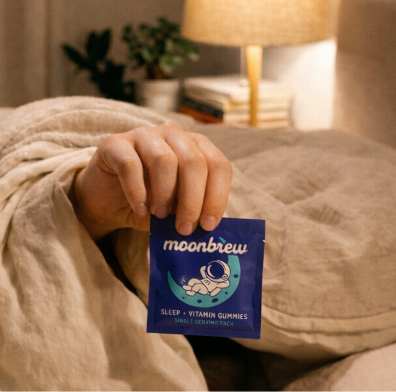 MoonBrew blue single-serve Sleep + Vitamin gummies packet with astronaut-on-moon graphic, held in a hand over a bed