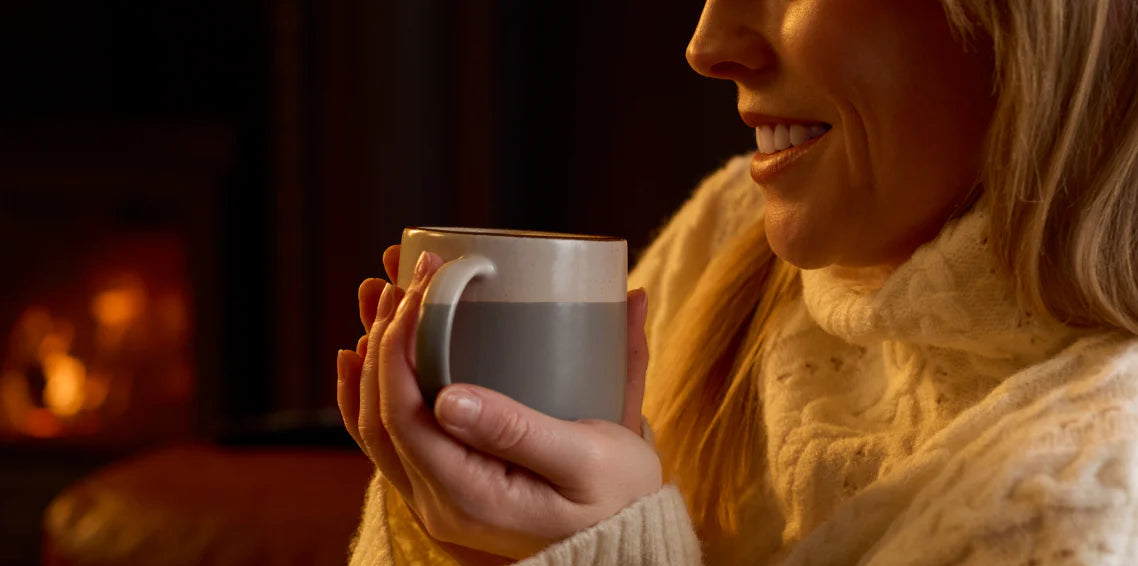 Woman smiling while holding a warm mug by a lit fireplace in a cozy sweater